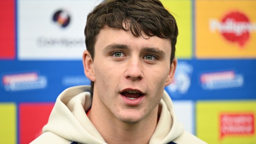 A young man with brown hair speaks to the media in front of a sponsorship sign