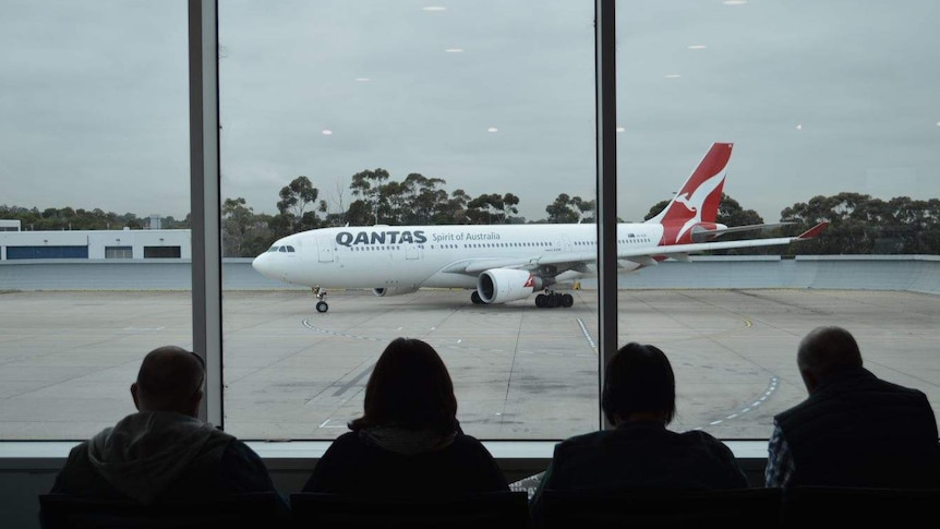 A Qantas plane taxies along a runway in overcast conditions while passengers seated inside watch on.