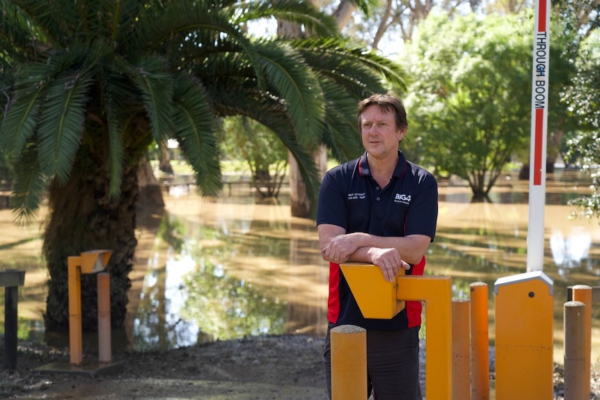 Matt Borrack leans on a yellow intercom panel as brown flood water covers the camping ground behind him.