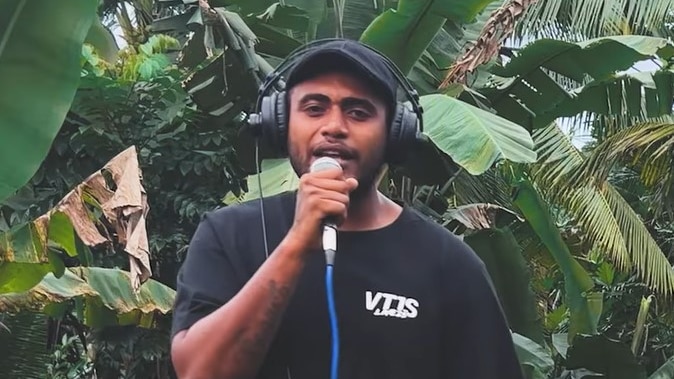 Man wearing black outfit and hat sings into a mic with headphones on standing infront of green lush outdoors.