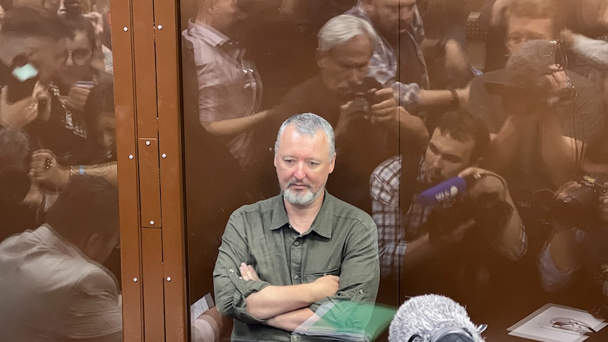 Igor Girkin sits with his arms folded in a glass box while people take photos 