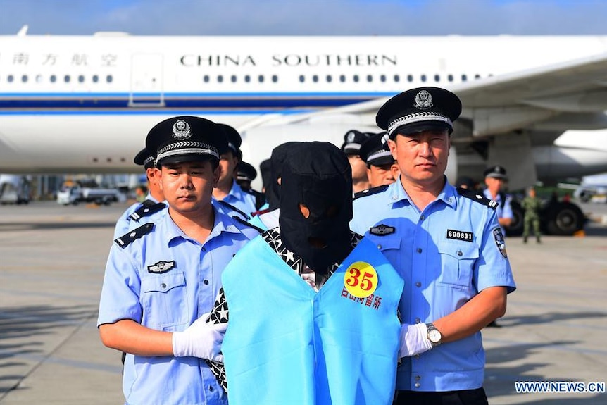 Chinese police stand with deported fraud suspects on the tarmac. The suspects are hooded and wearing numbered blue vests.