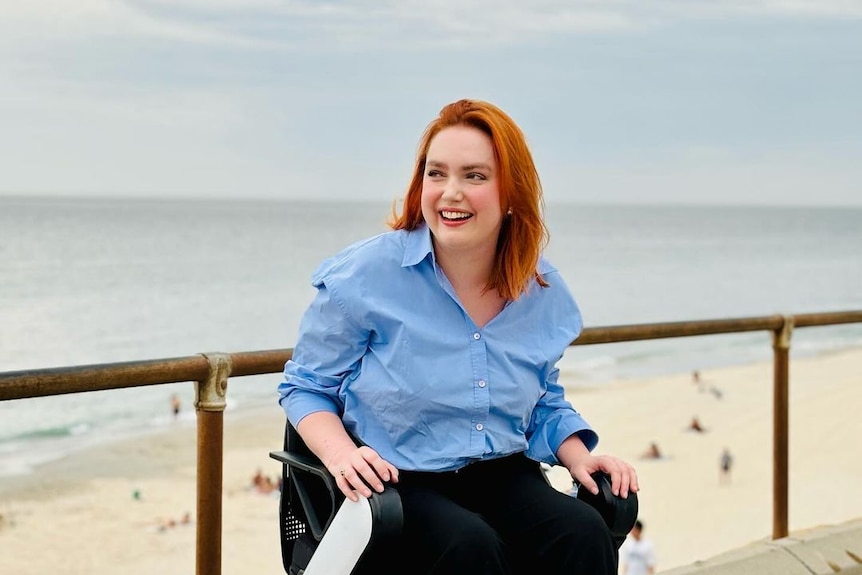 A woman with red short hair in a light blue shirt is in a wheelchair and laughing while in front of a beach outside 