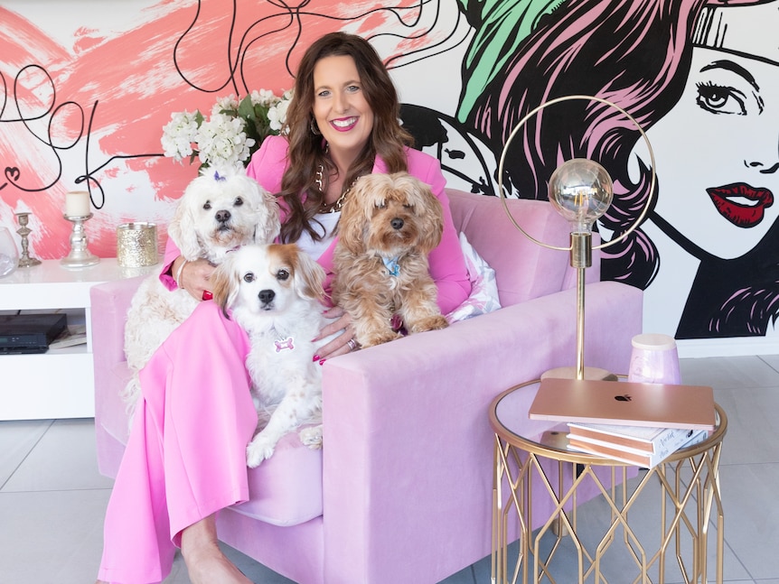 A women in a pink suits, sits on a pink armchair, holding three dogs. 