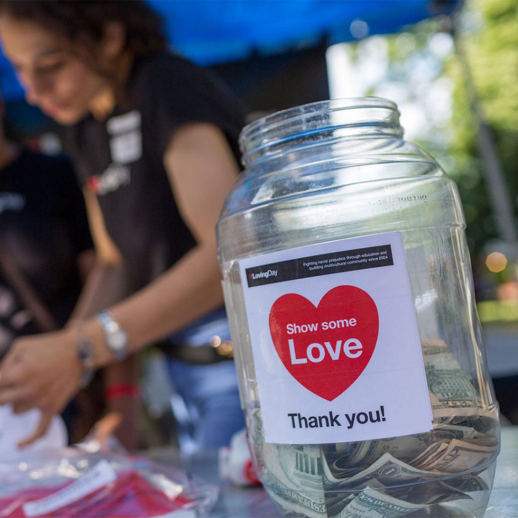 A transparent donation jar with some small bills inside and a label that says show some love in front of two volunteers at Loving Day NYC 2019.