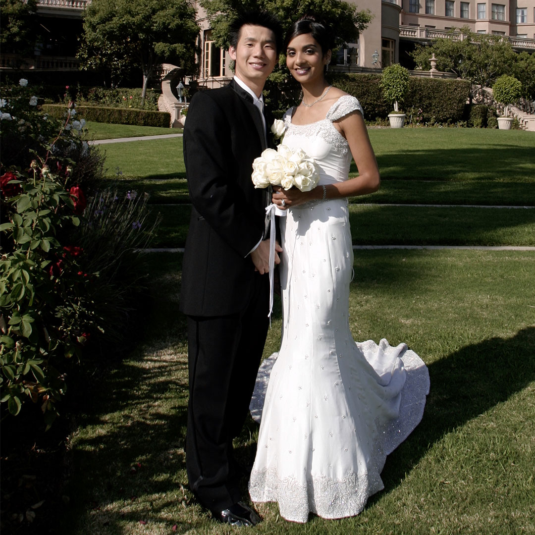 A wedding photo of a woman and a man smiling and standing outdoors on a grass outside of a large building.