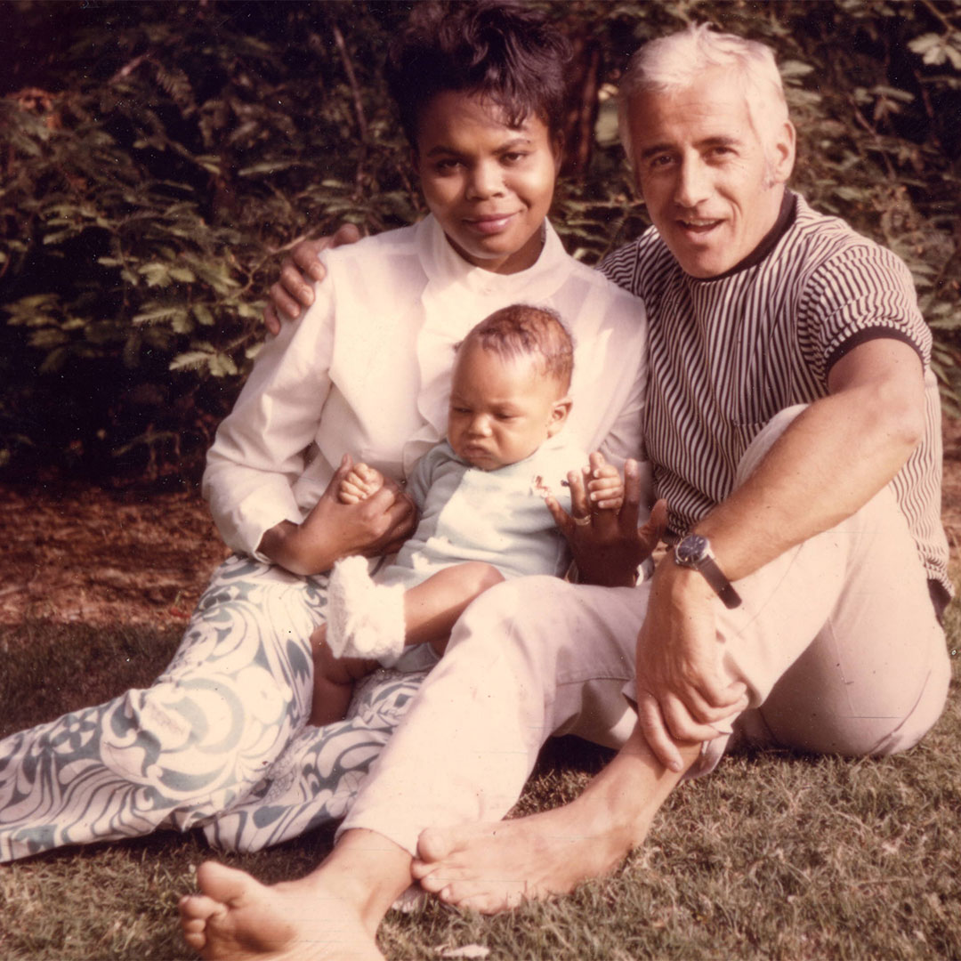A faded family photo of a woman and a man smiling with a baby sitting outdoors on the grass.