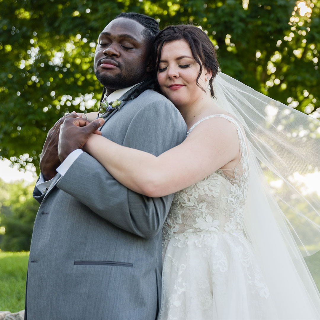 A wedding photo of a woman hugging a man from behind with their eyes closed, standing outdoors.