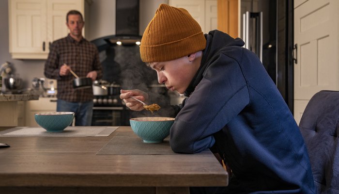 Teen boy sitting at a kitchen table eating soup served by his father