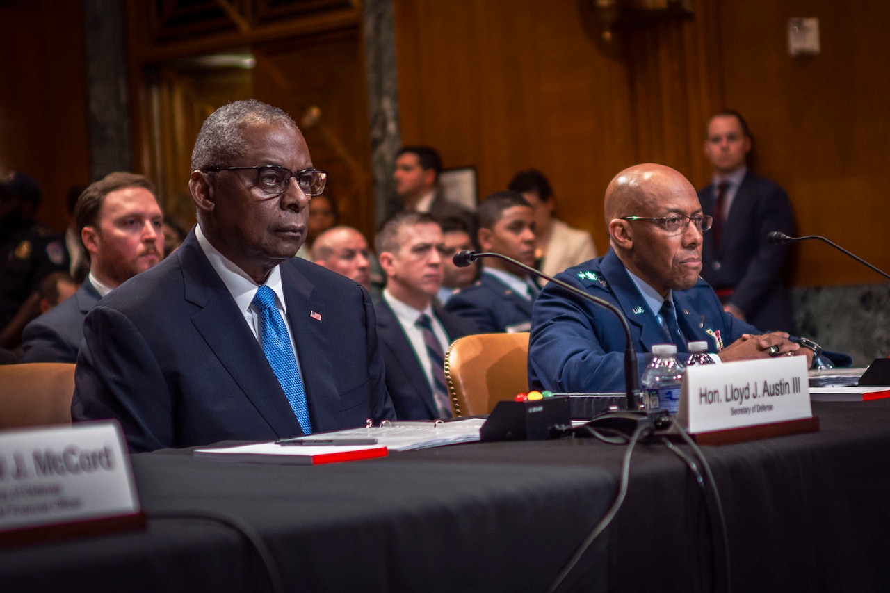Secretary of Defense Lloyd J. Austin III and Air Force Gen. CQ Brown, Jr., chairman of the Joint Chiefs of Staff sit behind a large table with others in the background.