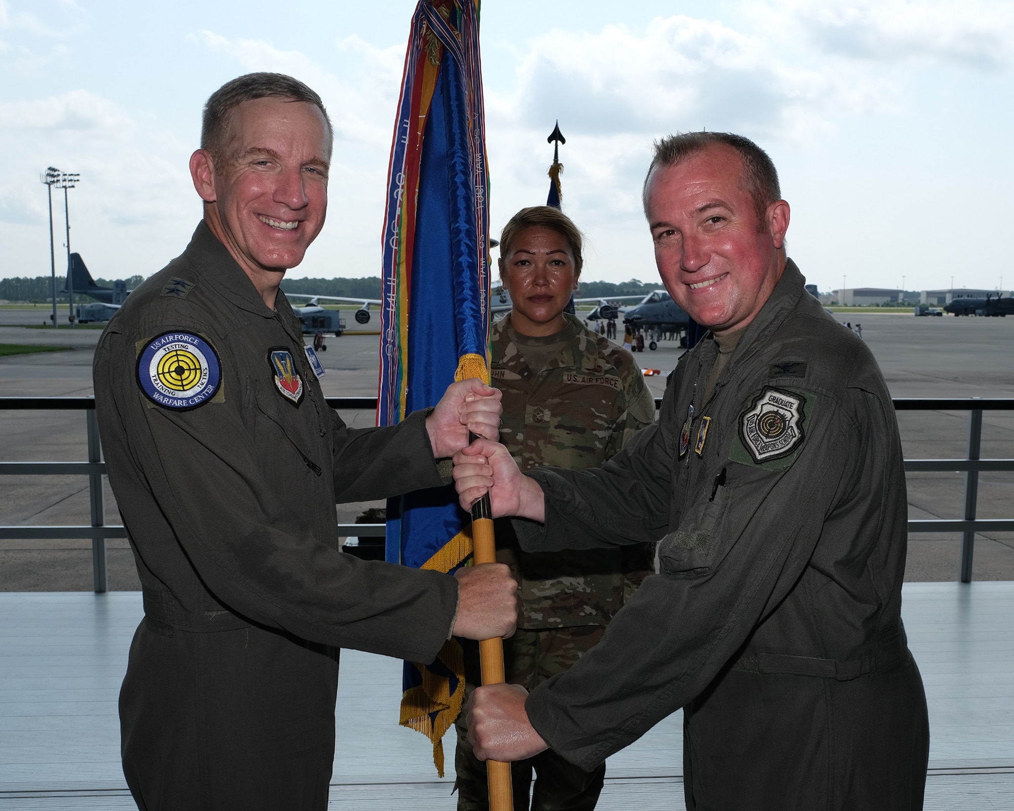 photo of three uniformed U.S. Air Force Airmen stand on a stand, two in the foreground holding a unit flag, while the third Airman stands at attention in the background are two A-10Cs and an airborne warning and control system, or AWACS, aircraft on Hurlburt Field flightline.