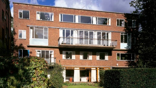 The garden front of 2 Willow Road. A square modernist building of four storeys stands central with green hedges and trees in the foreground.