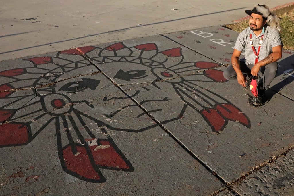 A picture of Native American advocate Carl Moore sitting next to Native American imagery painted along a walkway at a NY high school.