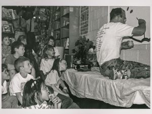 Black and white photo of man painting mural with his back to a row of seated children watching