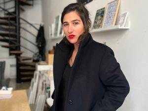 A woman wearing red lipstick and a black jacket poses in what looks to be an art gallery with spiral stairs