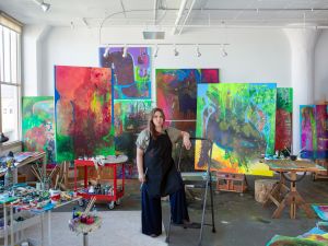 A woman wearing black overalls stands in a studio surrounded by large colorfui canvases