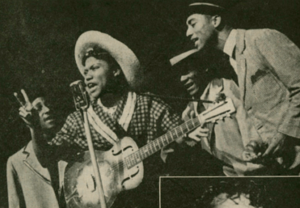 A black and white photograph of Sister Rosetta Tharpe performing in 1944. She is holding a guitar and singing into a microphone. A group of three men in suits sing behind her.