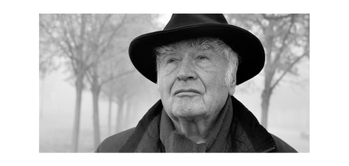 Martin Walser schaut in die Ferne, er trägt einen Mantel und einen Hut. Das Bild ist in schwarz-weiß. Im Hintergrund Bäume ohne Blätter.