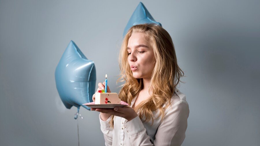 Девушка рядом с шариками дует на свечку в кусочке торта.