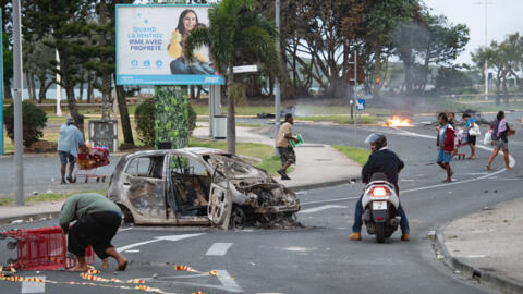Des personnes marchent dans la rue en poussant des chariots de supermarché à côté d'une voiture incendiée après le pillage d'un supermarché et le vandalisme de magasins dans le quartier de N'Gea à Nouméa, le 14 mai 2024.