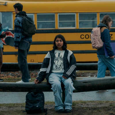 Vritika Gupta sitting in front of a school bus in a scene from 'Under the Bridge,' where she plays and an outcast teen who goes missing.