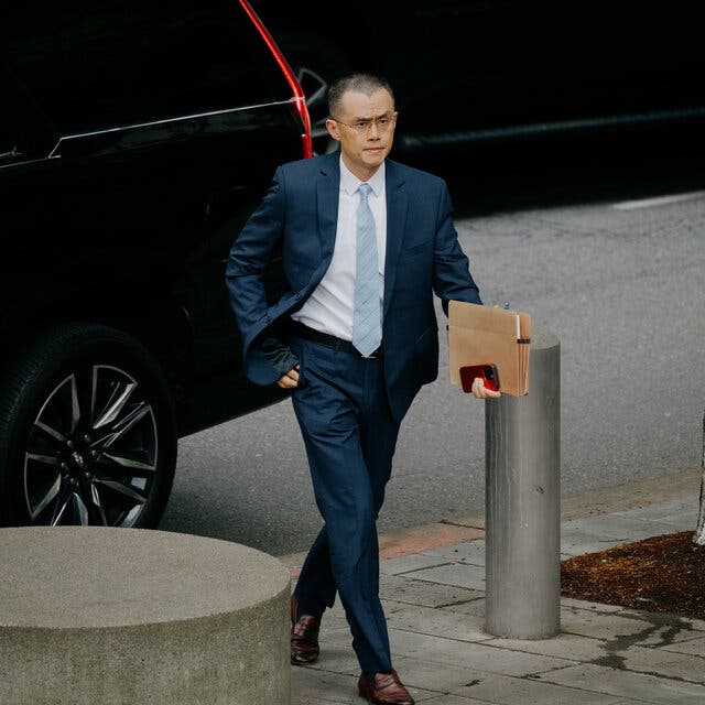 Changpeng Zhao walks away from a black S.U.V., carrying a file folder and a phone in his left hand. 