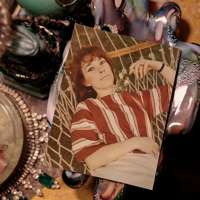 A color snapshot of Betty Gordon wearing a striped, red-and-white shirt and lying on a cot. The photograph is surrounded by jewelry.
