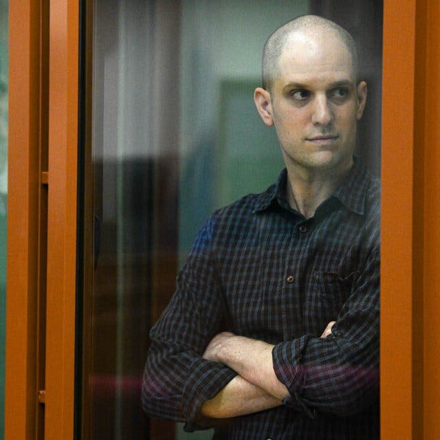 Evan Gershkovich, wearing a dark blue shirt, looked to his left while standing inside an orange-framed glass cage in a courtroom.